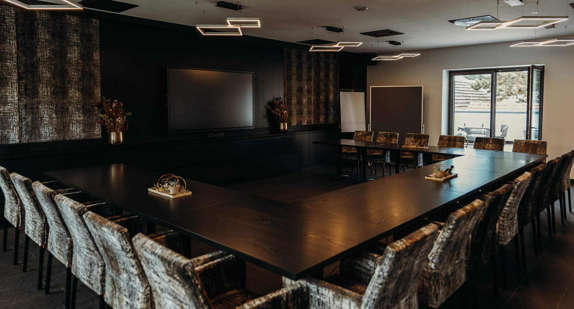 Moderner Konferenzraum mit U-förmigem Tisch, gemusterten Stühlen, TV, Flipchart und geometrischen Deckenleuchten.