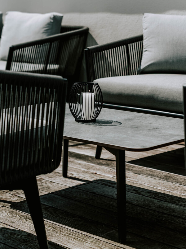 Eine weiße Kerze in einem schwarzen Drahthalter steht auf einem Tisch im Freien, umgeben von modernen Terrassenstühlen.