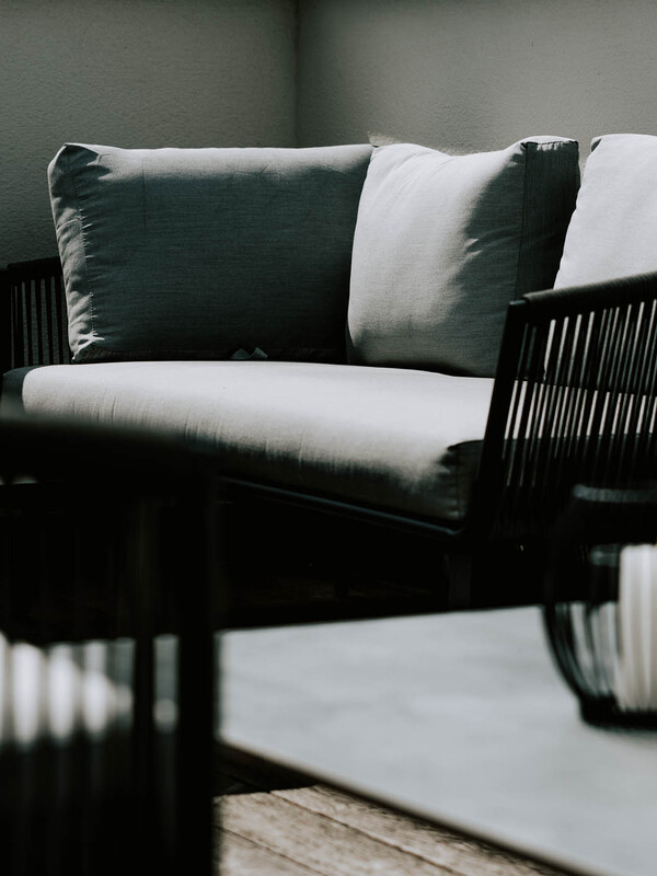 Graues gepolstertes Outdoor-Sofa mit schwarzem Rahmen im Sonnenlicht, neben einem Tisch mit unscharfen Objekten davor.