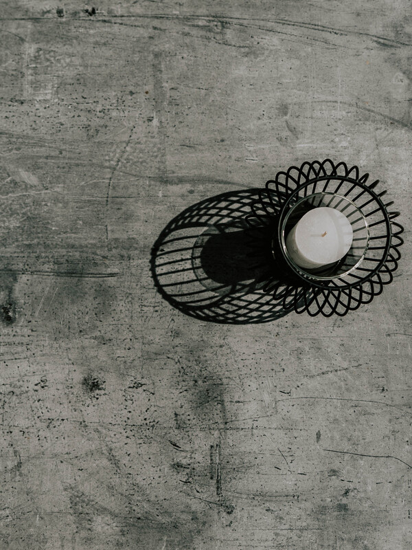 Eine kleine Kerze in einem Drahthalter wirft einen Schatten auf eine strukturierte Betonoberfläche.