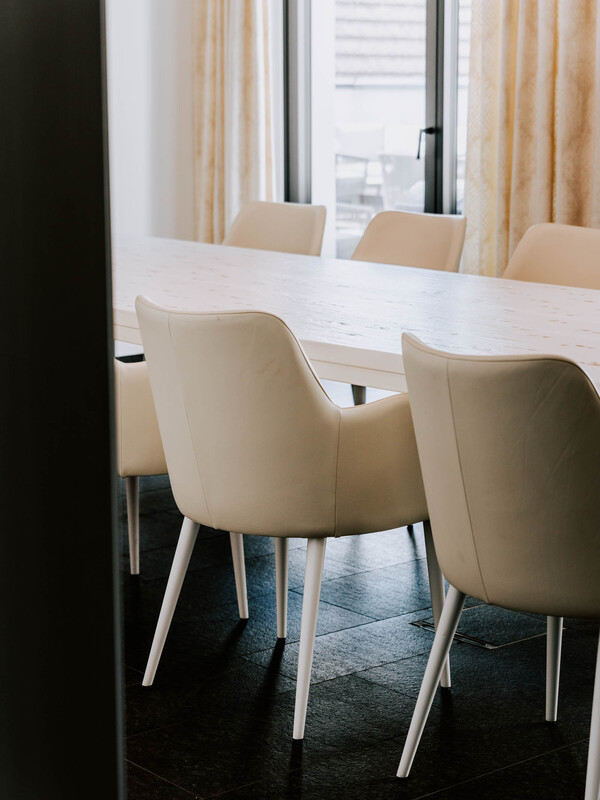Cremefarbene Stühle um einen langen weißen Tisch in einem modernen, sonnendurchfluteten Esszimmer mit großen Fenstern.