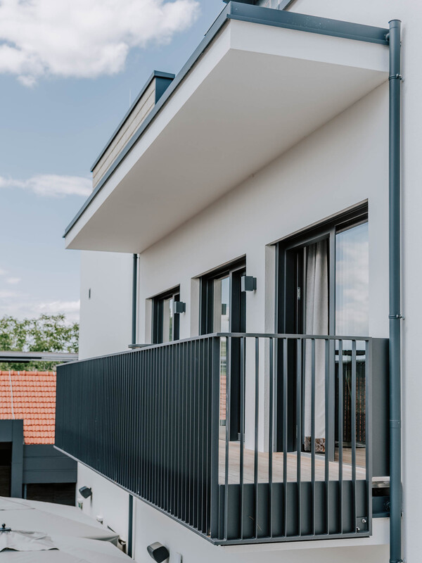 Moderner Gebäudebalkon mit Metallgeländer, großen Fenstern und einem Flachdach vor einem teilweise bewölkten Himmel.