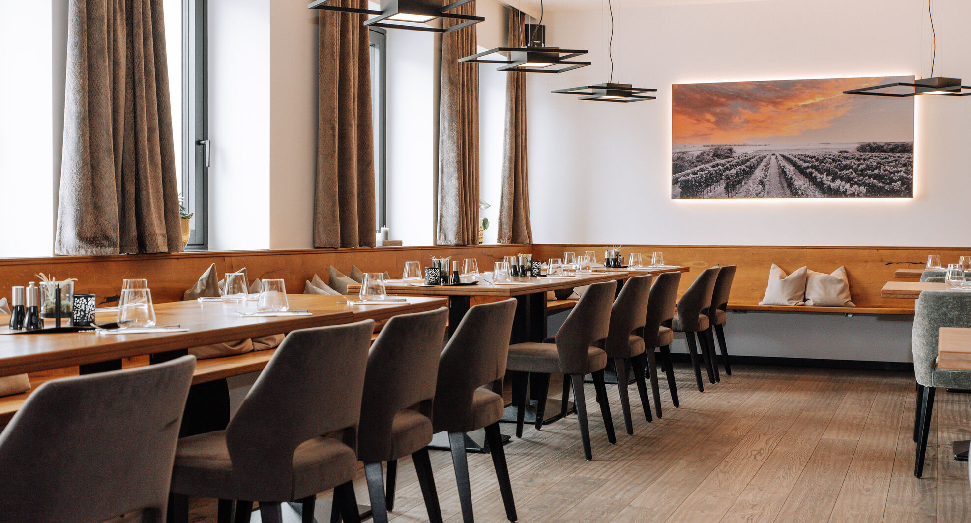 Modernes Restaurant mit Holztischen, grauen Stühlen, großen Fenstern und einem Landschaftsfoto an der Wand.