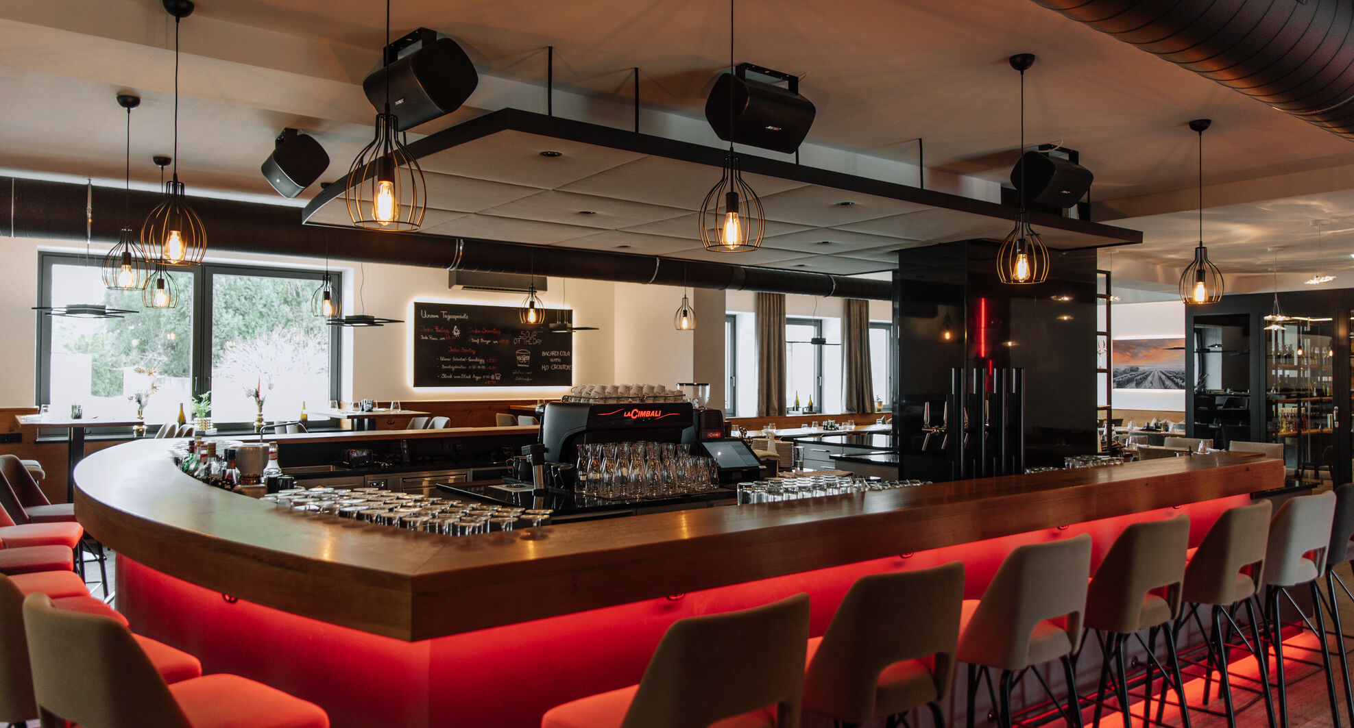 Moderne Bar mit geschwungenem Tresen, roter Beleuchtung, Barhockern, Hängelampen und großen Fenstern.