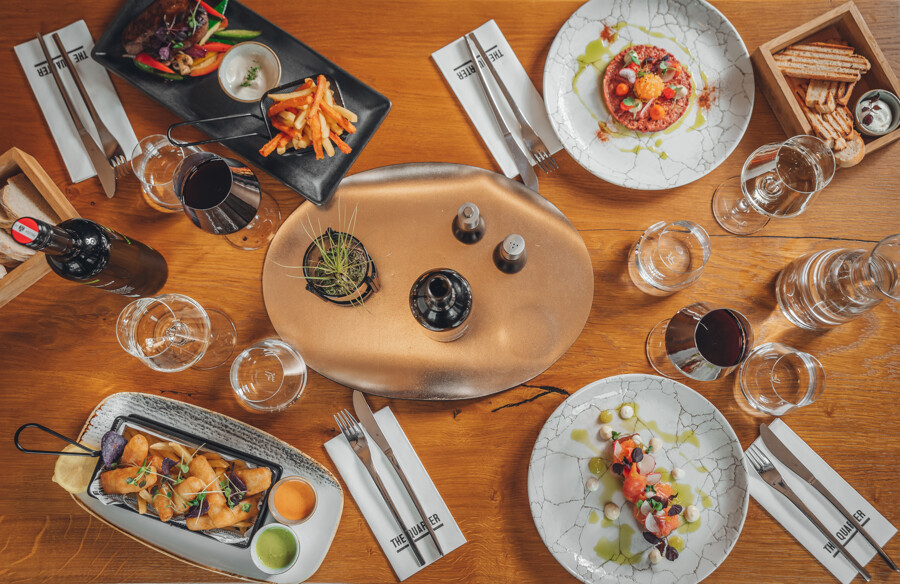 Ein Holztisch, gedeckt mit Gourmet-Häppchen, Besteck, Gläsern, einer Weinflasche und einem dekorativen Mittelstück.