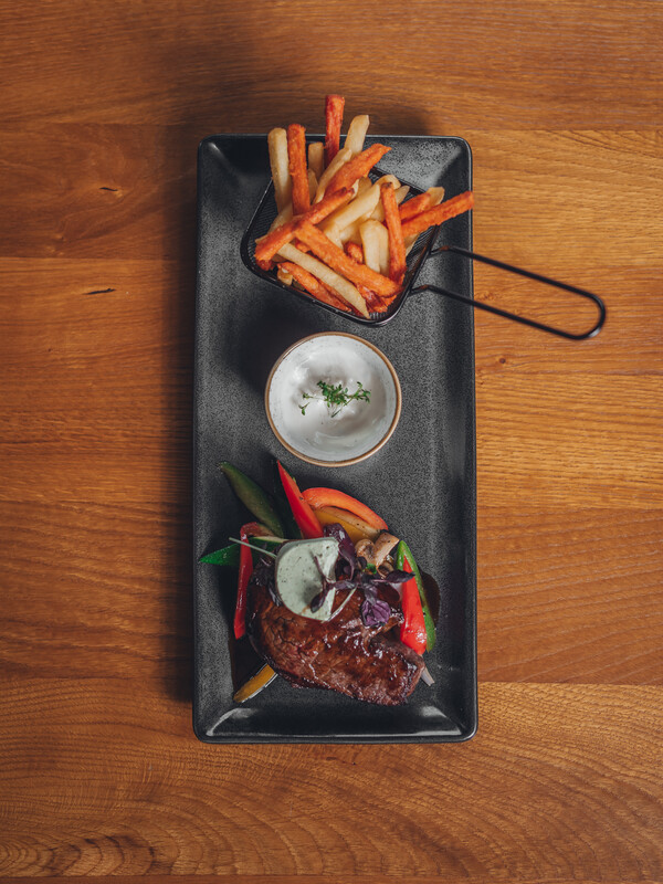 Rechteckiger Teller mit Steak, buntem Gemüse, Pommes frites in einem kleinen Korb und einem Becher mit Dip.