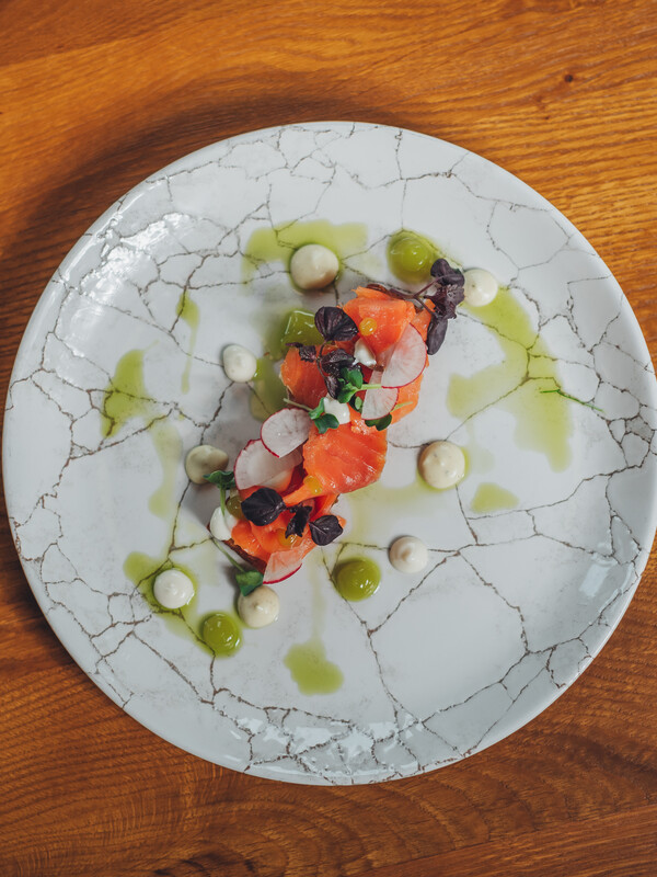 Ein Gourmet-Gericht mit geschnittenem Lachs, Kräutern, Radieschen und Soßentupfen auf einem weißen Strukturteller.