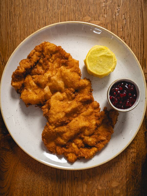 Zwei panierte Schnitzel mit einer Zitronenspalte und einer kleinen Tasse Preiselbeersauce auf einem weißen Teller.